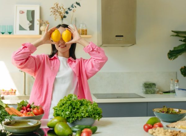 A woman playfully holding lemons over her eyes in a bright, lively kitchen setting filled with fresh vegetables.